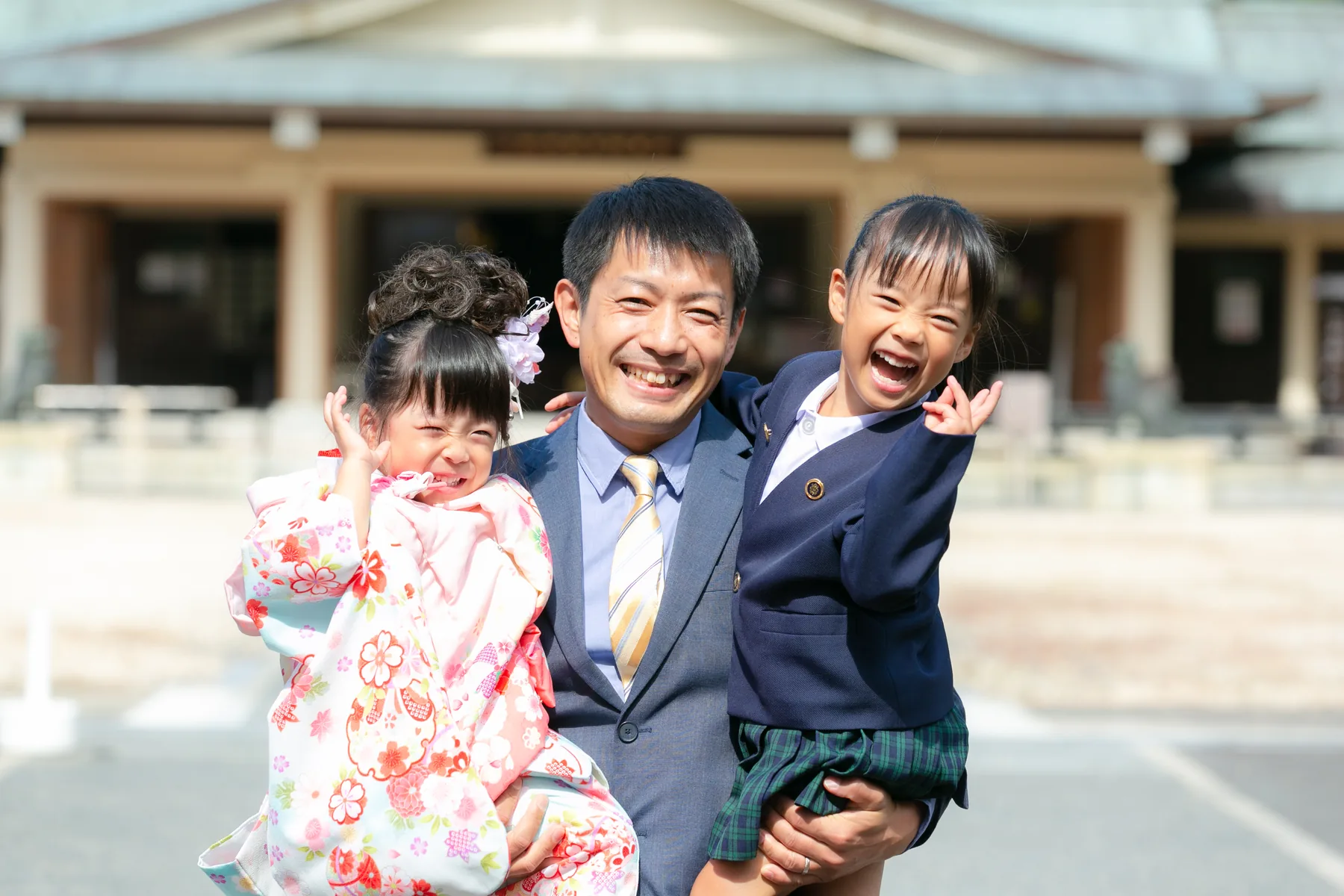 神社の前で、七五三の着物と袴を着た子どもふたりを抱き上げるお父さん