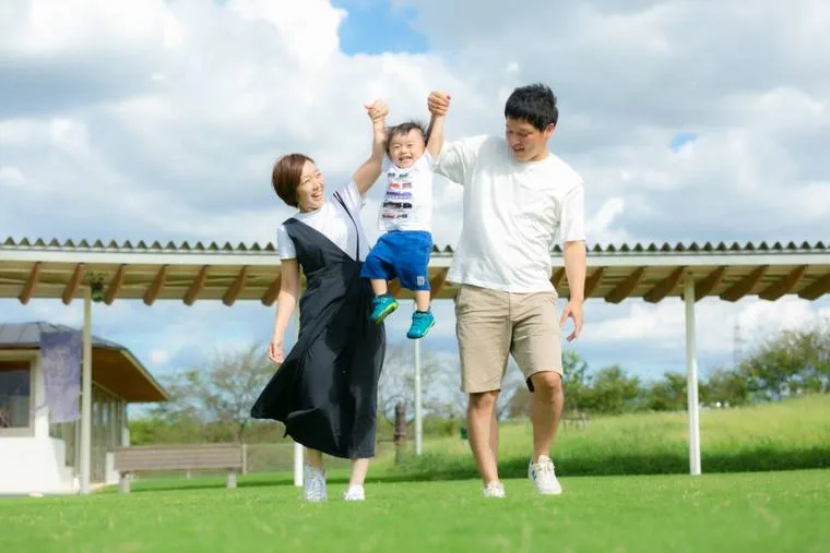 芝生の上で、お父さんに抱き上げられて笑う子どもと、隣で手を上げるお母さん