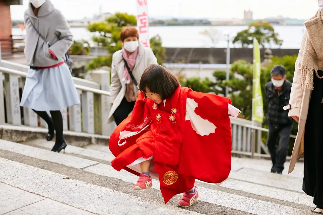赤い被布を着た女の子が、神社の階段を勢いよく駆け上がっている。まわりを参拝の人が歩いている