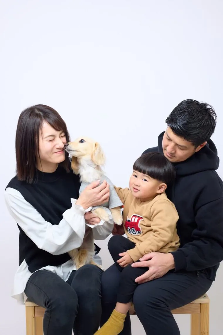 スタジオで、小さな犬を抱く母親と、父親に抱かれた子どもが並んでいる家族写真