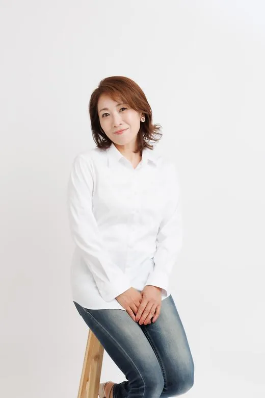 白い背景の前で、椅子に腰かけてこちらを見ている白いシャツの女性のプロフィール写真