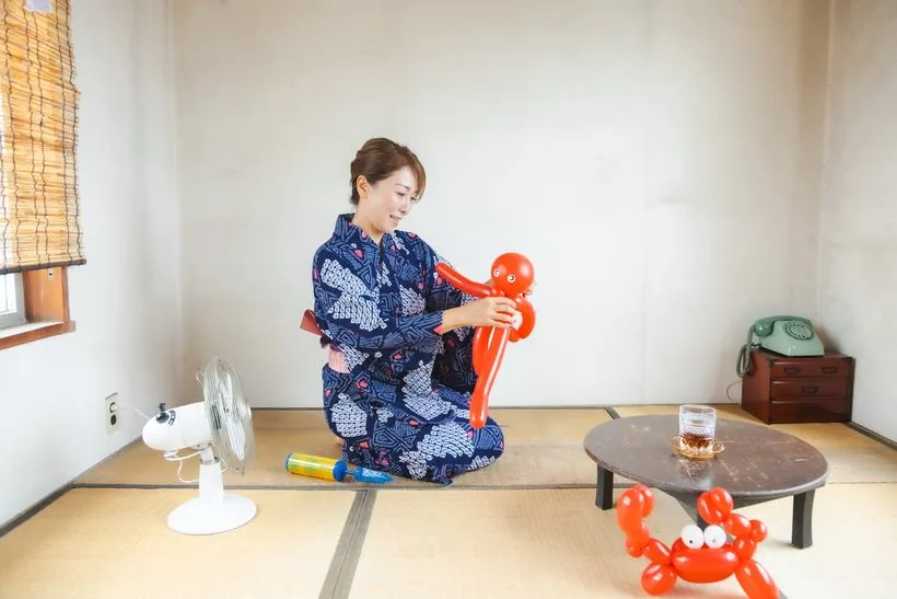 スタジオの和室で、浴衣を着てバルーンアートを作っている女性。扇風機や黒電話が置かれている