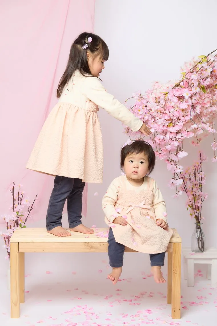 ベンチの上に立つ女の子が、桜の枝から花びらを摘んで、隣に座る妹の頭の上に降らせている