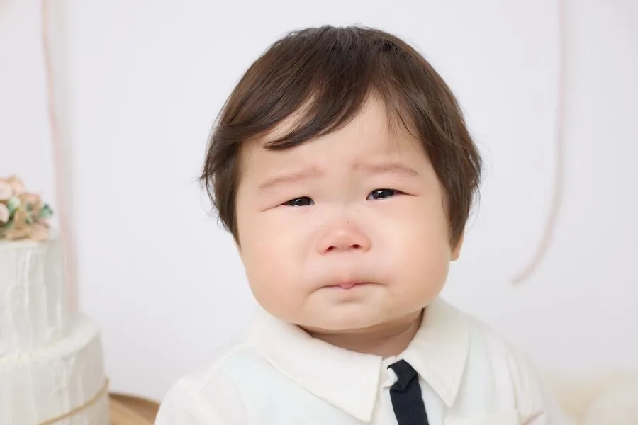白い背景の前で、眉を寄せて今にも泣き出しそうな顔をしている赤ちゃんの表情。左端にケーキが写っている