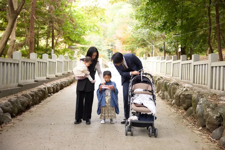 紅葉の参道で、もらったお菓子を覗きこむ家族。父親と母親と着物姿の男の子が、それぞれ手元のお菓子に目を向けている